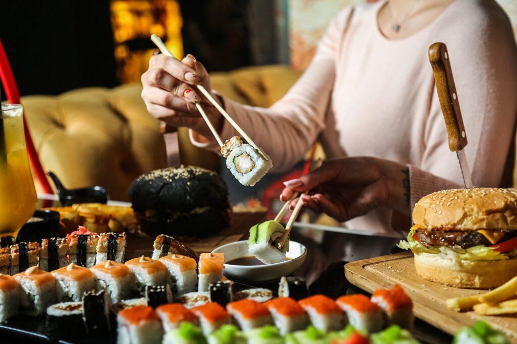 side view woman eats mix sushi rolls with soy sauce and burgers on the table
